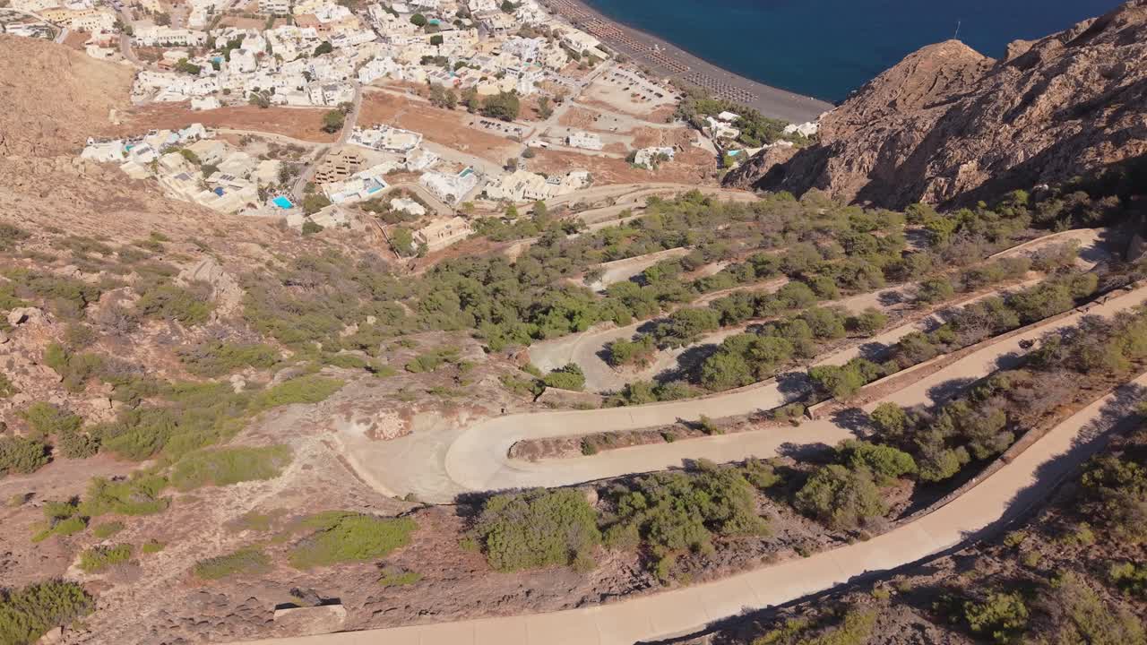 Kamari, Santorini'nin panoramik drone görüntüsü. Çekim, kayalıklara oyulmuş ünlü dağ zikzak yolunu, geniş siyah kumlu plajı ve masmavi Ege Denizi'ni gözler önüne seriyor.
