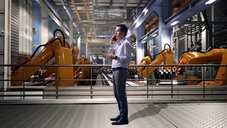 full body side view of an asian male professional worker standing with industrial robotic arms working, he is speaking with someone in the phone and moving his left hand