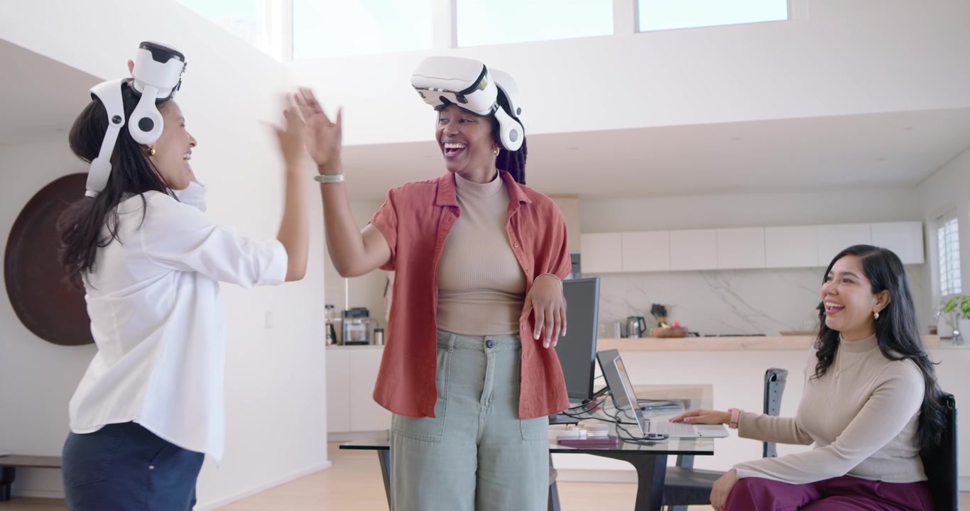Diverse colleghe seguono le indicazioni di una di loro, usando visori VR e battendosi il cinque in un ufficio domestico.