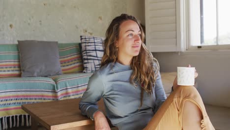 caucasian woman sitting on floor in living room drinking coffee, looking out of window and smiling