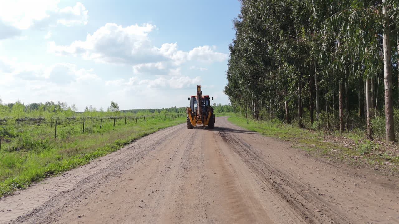 Aerial rearview tracking, tractor driving on a dirt road by green field, traveling to work site, surrounded by open farmland