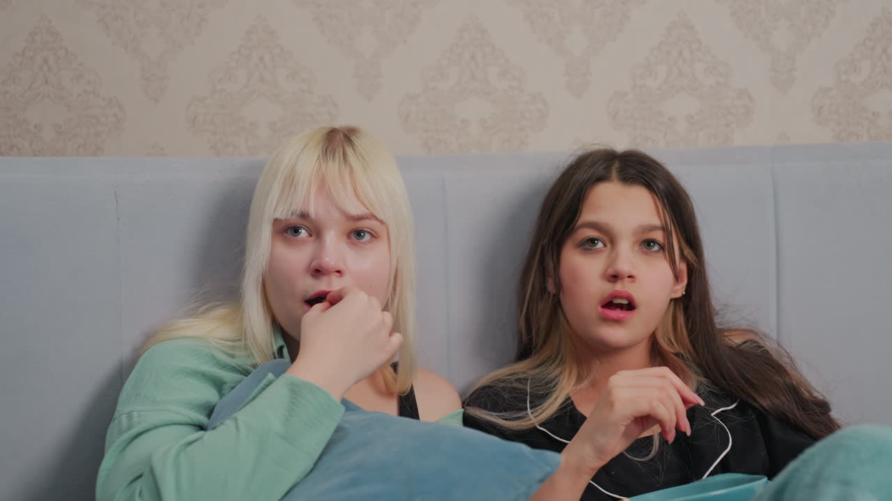 Two young siblings sit on bed with focused expressions while watching movie, holding popcorn in hands and wrapped in soft blankets, portraying immersive viewing moment in calm bedroom