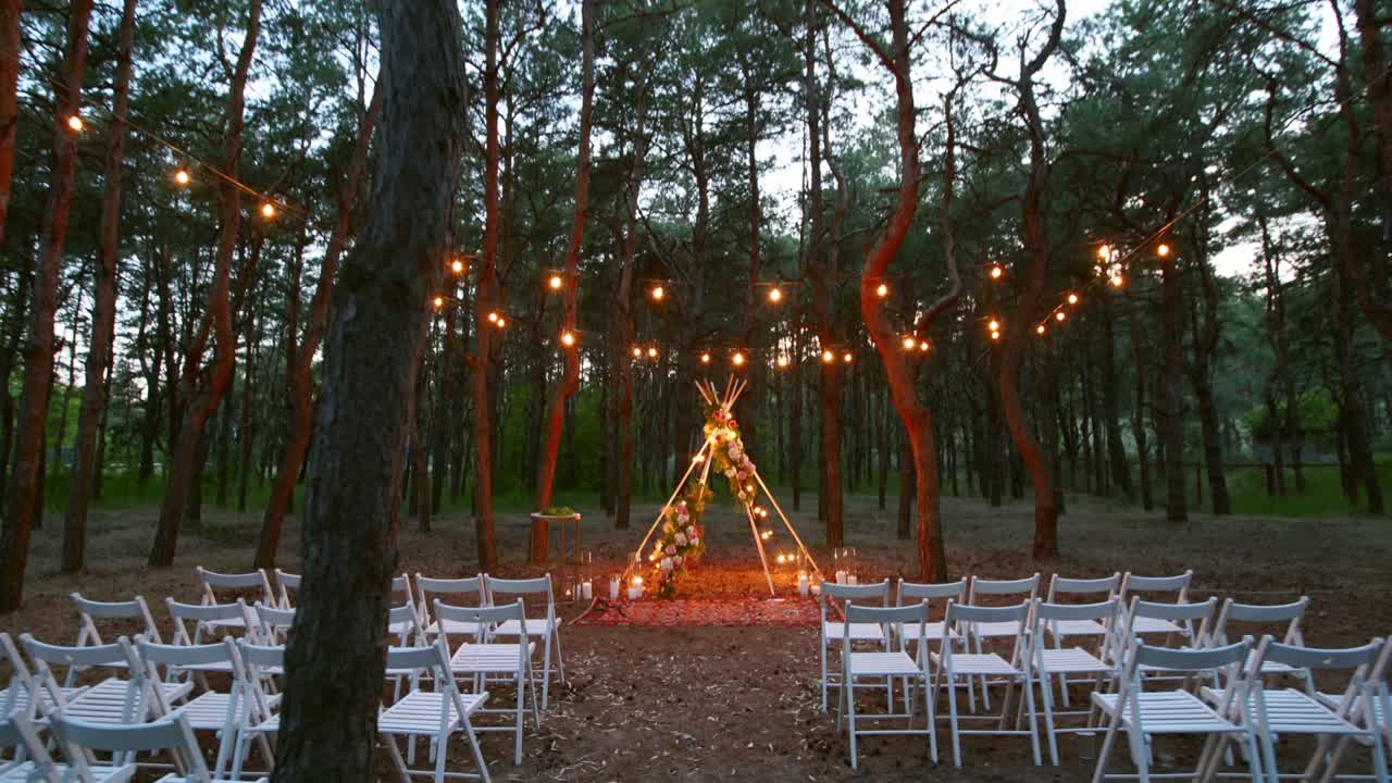 Festive string lights illumination on boho tipi arch decor on outdoor wedding ceremony venue in pine forest at night. Vintage string lights bulb garlands shining above chairs at summer rural wedding