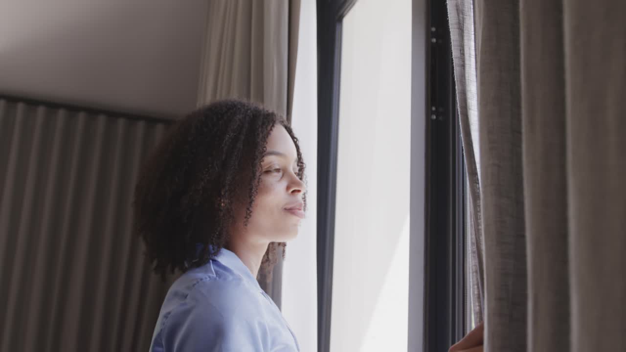 African American woman in blue top noticing dawn pulling drape at window letting daylight into room