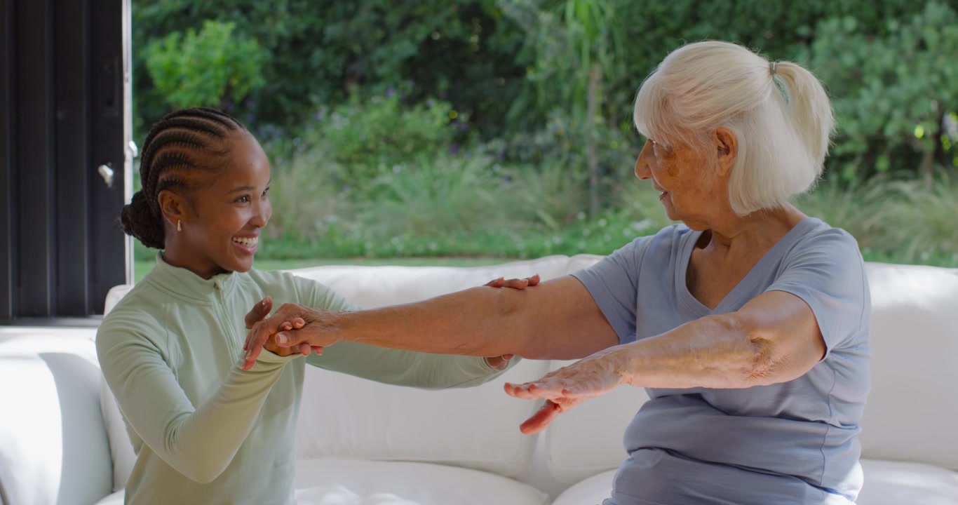 Female caregiver guiding senior woman starting by taking wrist, steadying arm for mobility on sofa