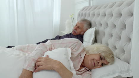 senior grandparents couple lying and sleeping in bed. woman getting disturbed with man snoring