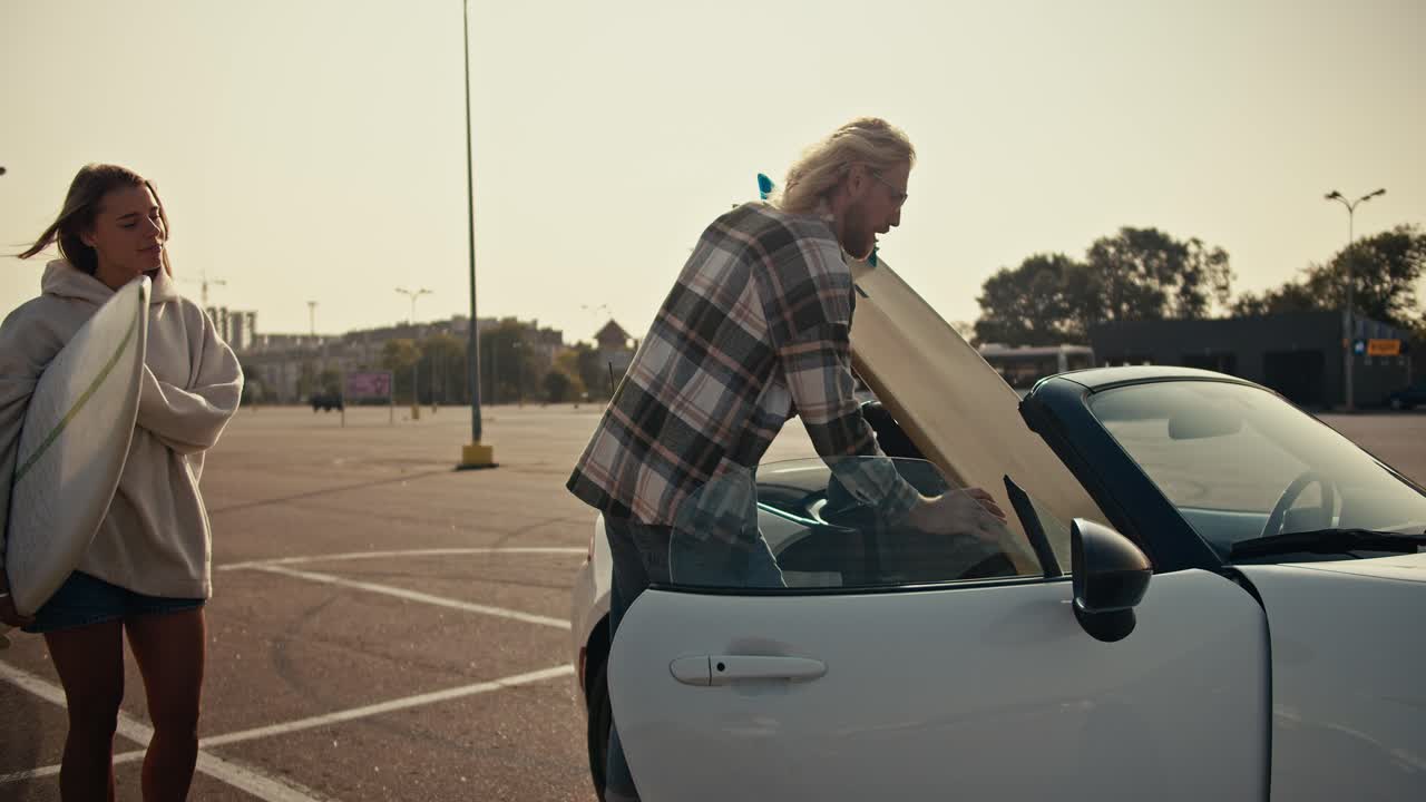 안경과 카레드 셔츠를 입은 금발의 남자가 그의 서프보드와 그의 금발 여자친구의 보드를 색 컨버터블에 넣습니다. 행복한 커플이 도시에서 서핑을 한 후 집으로 운전할 준비를 하고 있습니다.