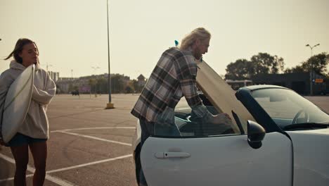 A-blond-guy-in-glasses-and-a-plaid-shirt-puts-his-surfboard-in-a-white-convertible-and-his-blonde-girlfriend's-board-too.-Happy-couple-getting-ready-to-drive-home-after-surfing-in-the-city