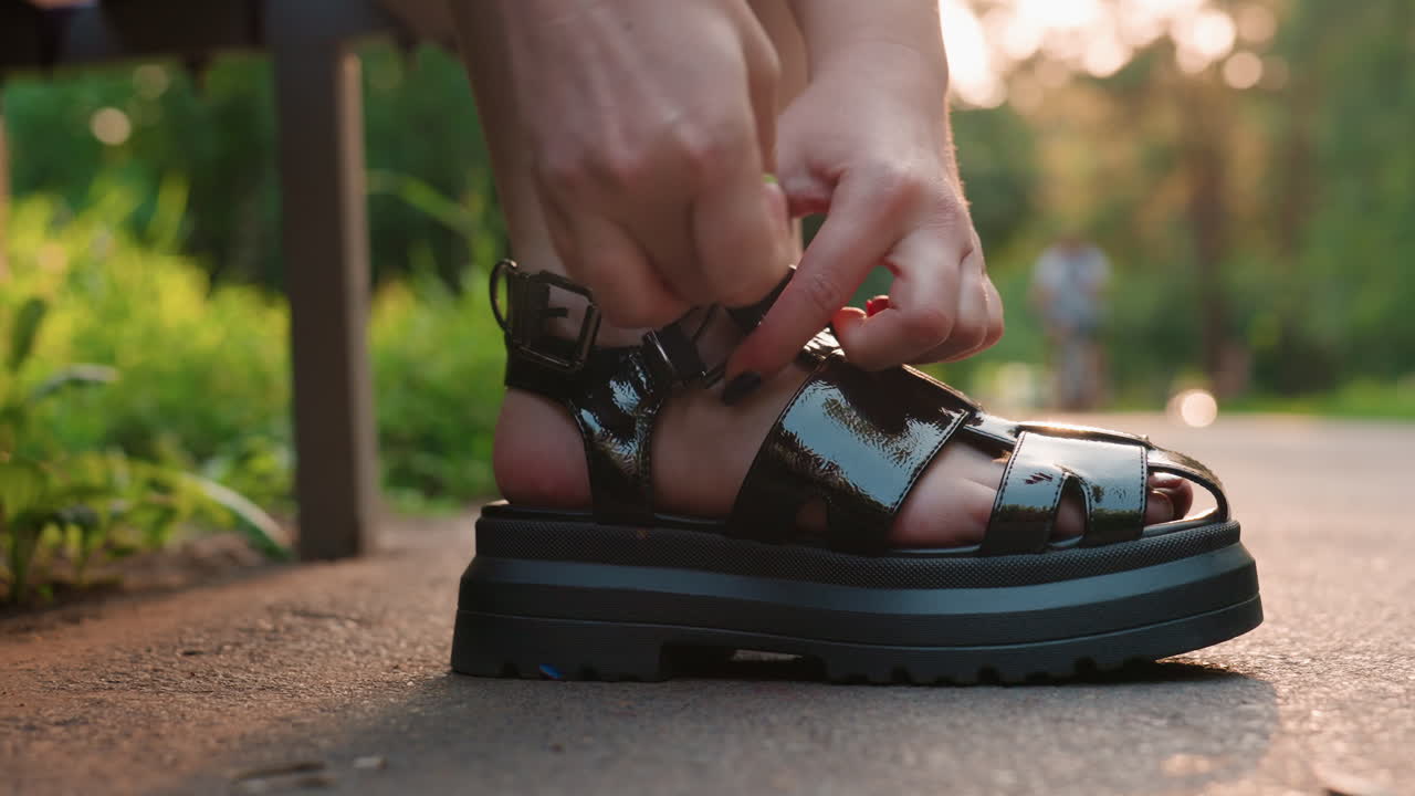 Estilista de moda ajustando un zapato en un parque, esteticista profesional ayudando con el calzado en un banco del parque, estilista experto ajustando cuidadosamente la correa de una sandalia a una modelo con un exuberante fondo de parque