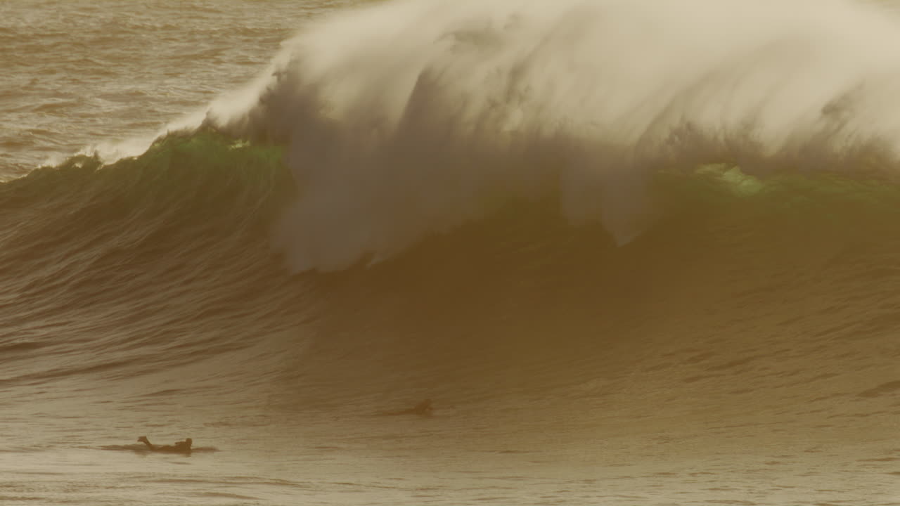 Clean face of wave curls with rich golden hue as mist spray picks up over top, surfers dive below