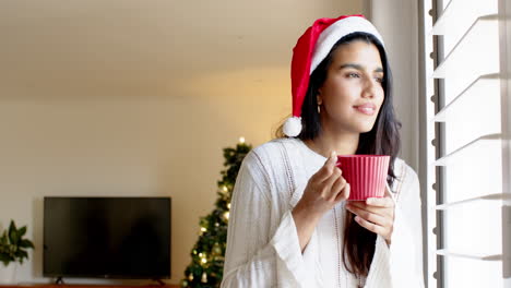 christmas time, indian woman in santa hat enjoying hot drink by window at home