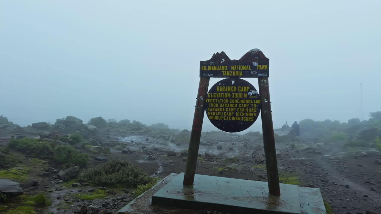 Panning shot shows Baranco Camp sign on a foggy alpine plateau at Mount Kilimanjaro, with a yellow tent and rugged rock wall in the background