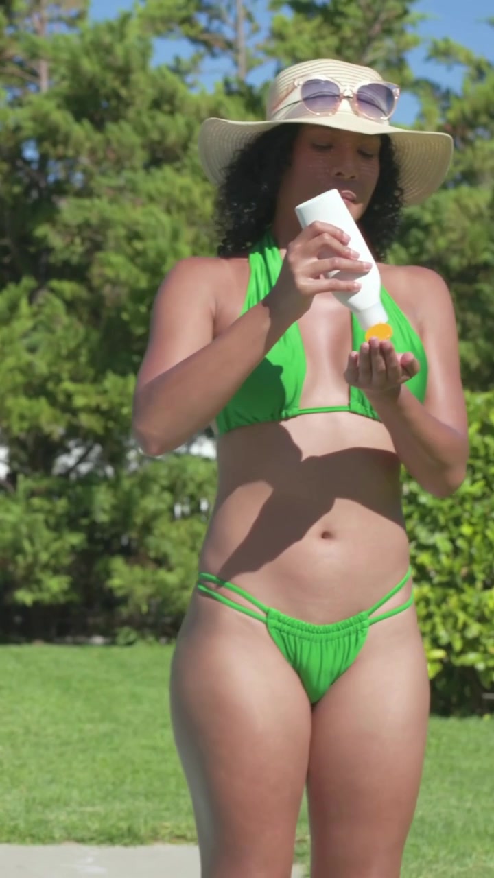 Vertical video: Walking into frame woman applying sunscreen at pool to avoid sunburn, green bikini