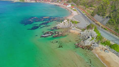 sweeping aerial shot pulling back over turquoise water and rocky shores to reveal the picturesque uradome coast, a seaside village, and a coastal road in tottori, japan