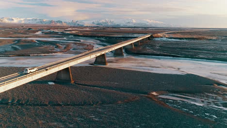 vehicle travelling across reynisfjara main ring road over black sand beach towards snow capped icelandic mountain range, rising aerial view