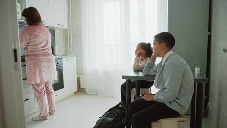 mother in pink velour pajamas preparing breakfast at kitchen countertop while children in school uniforms sit at table waiting with backpacks near window bright morning home routine scene