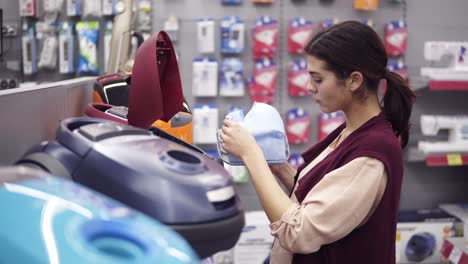 Female-costumer-choosing-vacuum-cleaner-in-appliance-store.-Coming-up-to-a-showcase-row-taking-a-dustbag-out-of-vacuum-cleaner-and-examining-it-carefuly.-Looking-for-domestic-equipment.