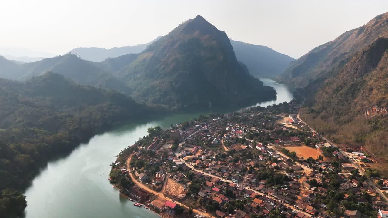 Drone riprende il villaggio di Nong Khiaw adagiato lungo il fiume Nam Ou nel Laos settentrionale, circondato da rigogliose montagne carsiche, con il tempio buddista Wat Sop Houn visibile tra la fitta vegetazione.
