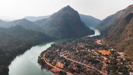 drone captures nong khiaw village nestled along the nam ou river in northern laos, surrounded by lush karst mountains, with the buddhist wat sop houn temple visible among dense foliage