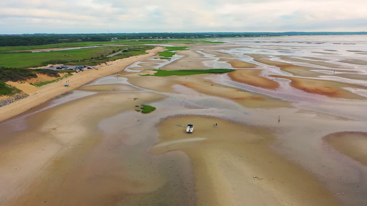 Aerial view of expansive sand flats at low tide where a stranded boat sits among tide pools, sculpted ridges, and patches of green marsh grass stretching across the coastal landscape