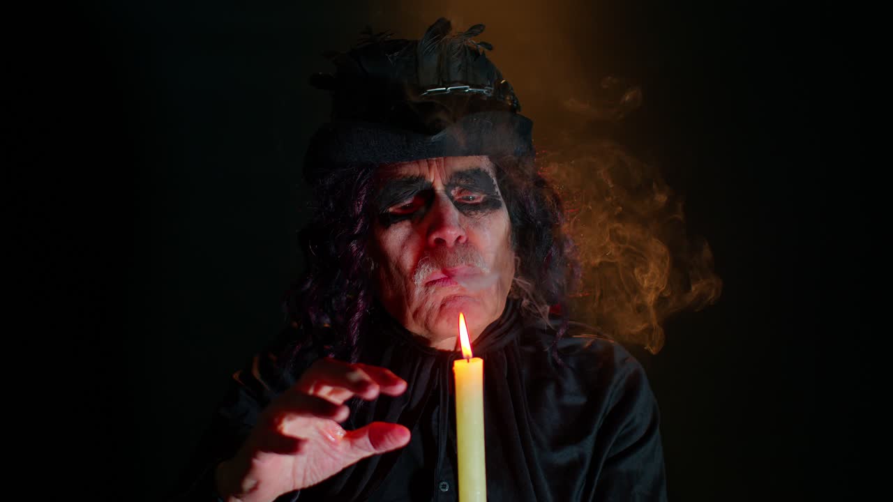 Sinister man with scary Halloween witcher makeup in costume making voodoo magic rituals with candle