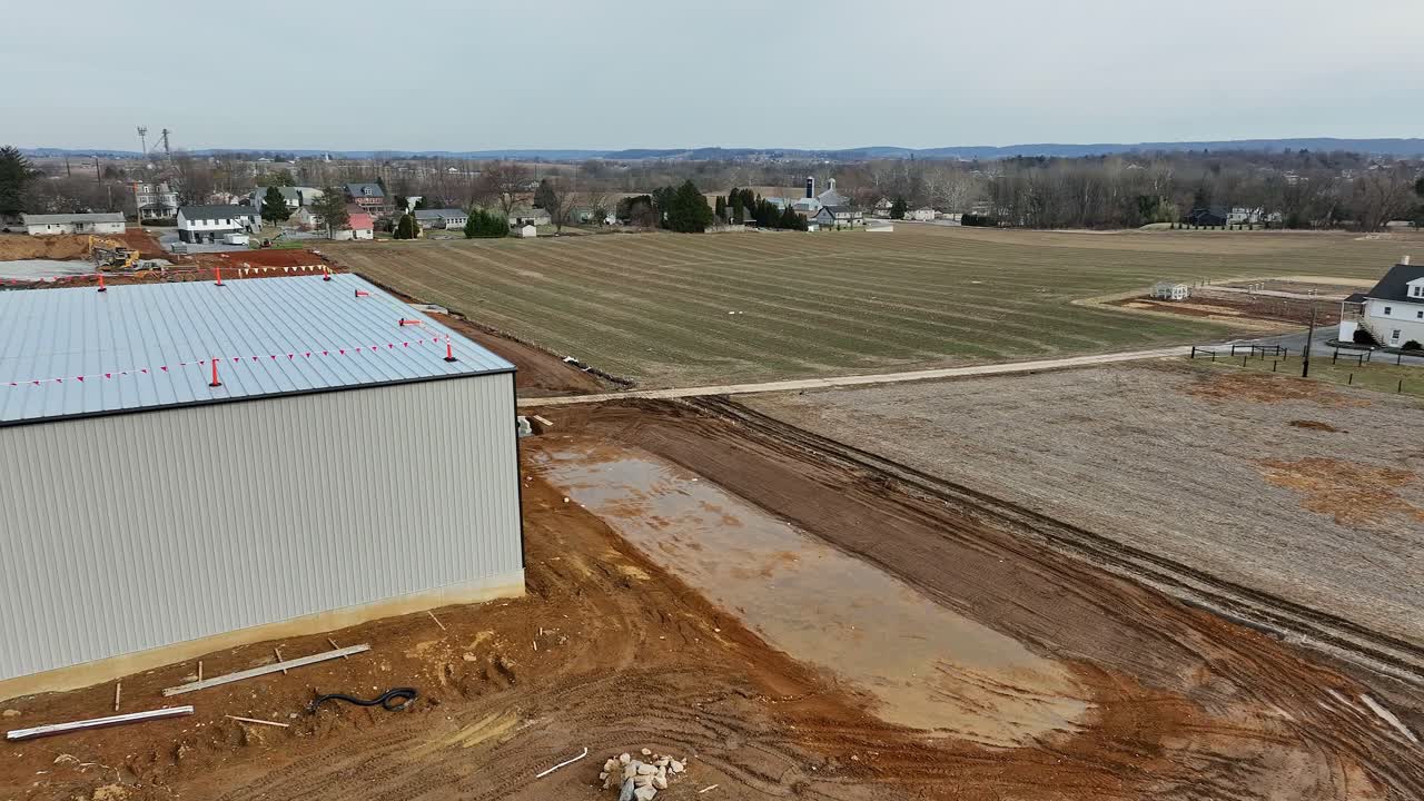A construction site is actively developing, with land being prepared for building. The view captures neighboring fields and structures nearby, highlighting ongoing work in the rural area.