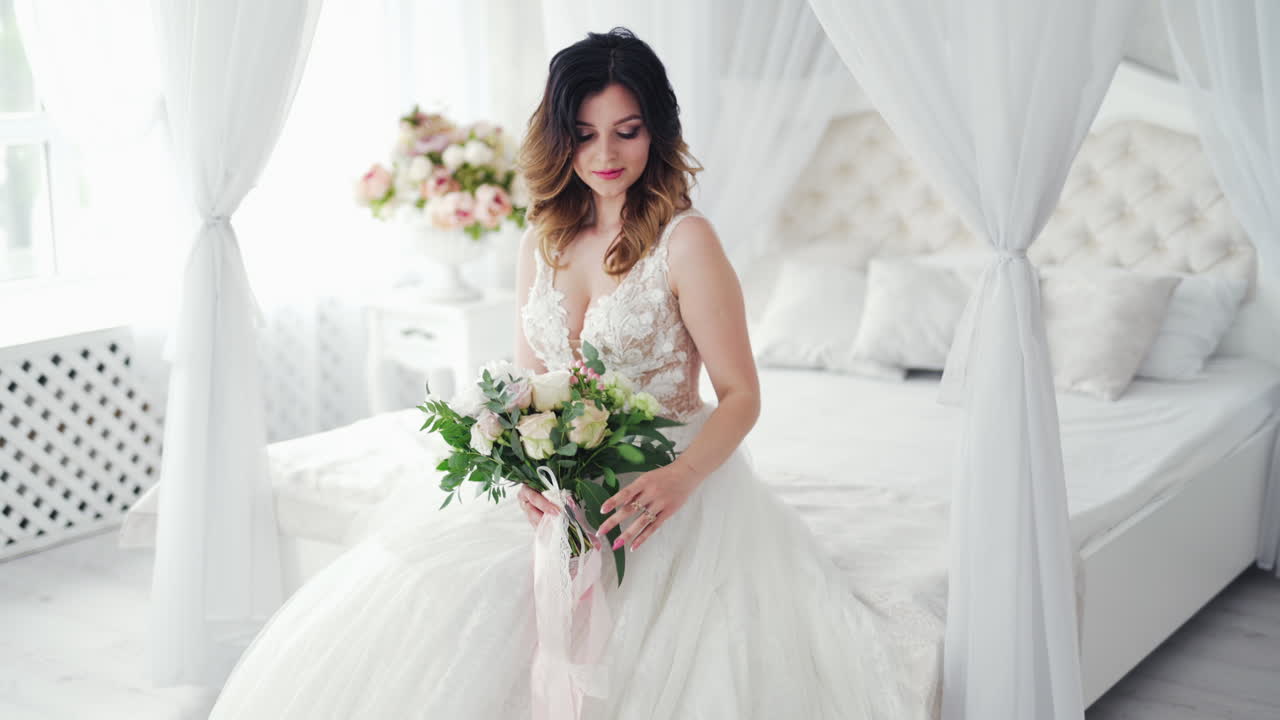 Video shot of a young bride in a wedding dress on the background of a bed. Attractive model with modern hairstyle in white dress is posing to camera inside the light room.