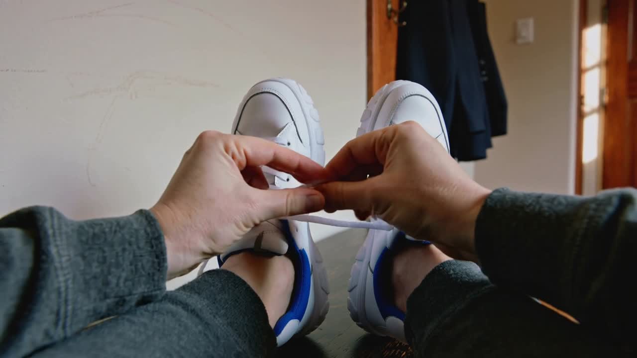 Un adulto si accorge di un laccio slacciato, si china per allacciare delle scarpe da ginnastica bianche all'ingresso, indossando una tuta grigia