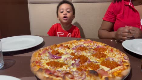 adorable niño de dos años impaciente por comer su primera pizza dentro de un restaurante, sentado junto a mamá