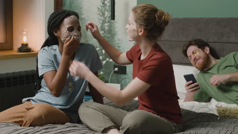 and girl applying facial mask sitting on the bed while their male roommate are using smartphone and laughing