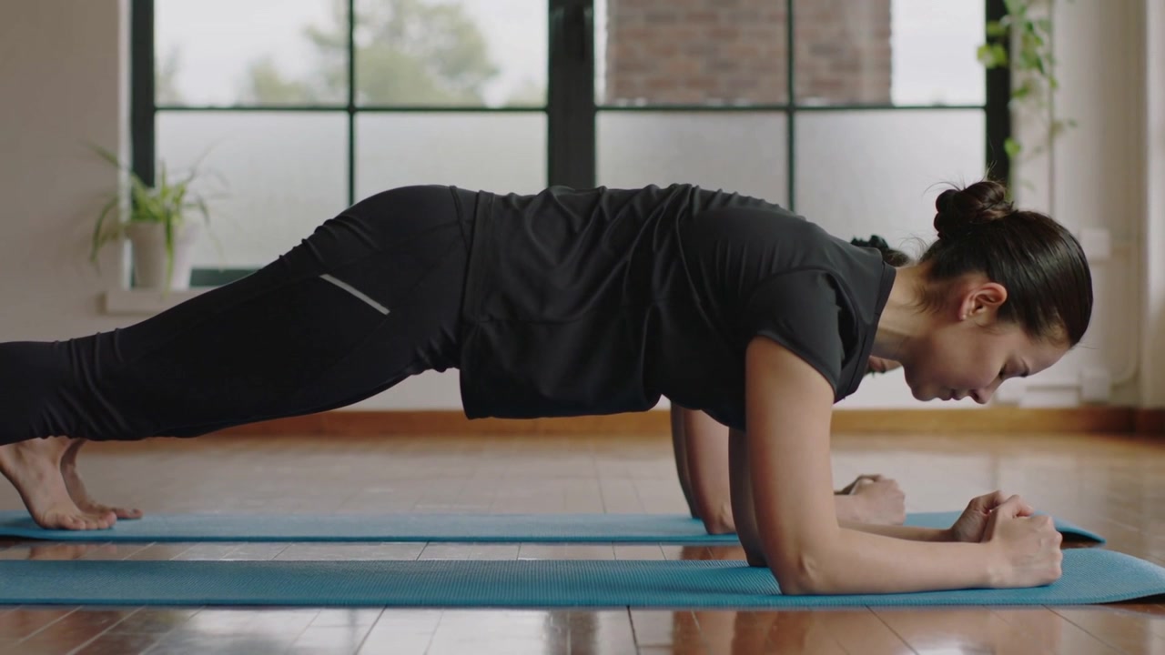 Wanita dengan bahu bertumpuk mengenakan perlengkapan hitam melakukan plank lengan bawah di atas matras biru dekat jendela, melatih otot inti