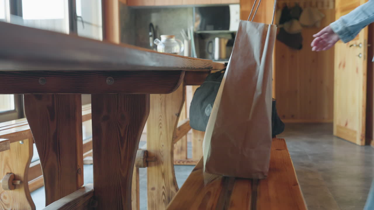 Hand in denim sleeve reaching for paper shopping bag and black duffel resting on wooden bench inside rustic kitchen with warm wood paneling and natural lighting filtering through nearby windows