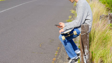 Side-view-of-cool-young-caucasian-man-with-skateboard-using-mobile-phone-at-countryside-road-4k