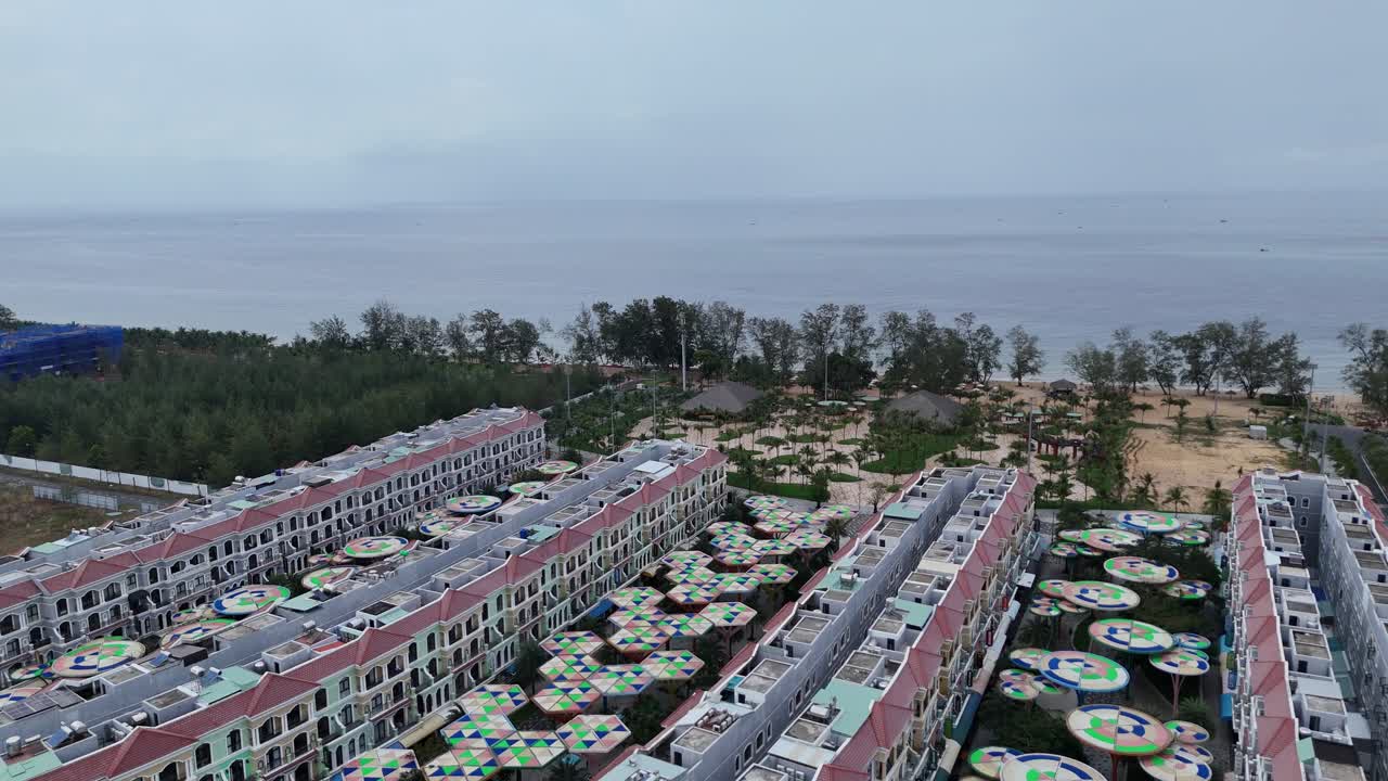 This aerial shot shows a large development of multi-story buildings situated near a coastline. The buildings appear uniform in style, with red-tiled roofs and light-colored facades.colorful structures