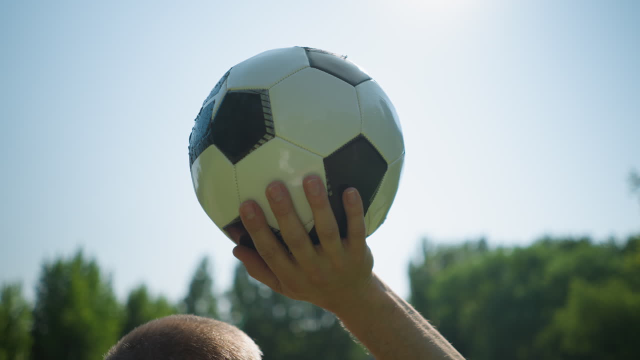 Close-up of a middle-aged man holding a soccer ball high above his head against a clear blue sky