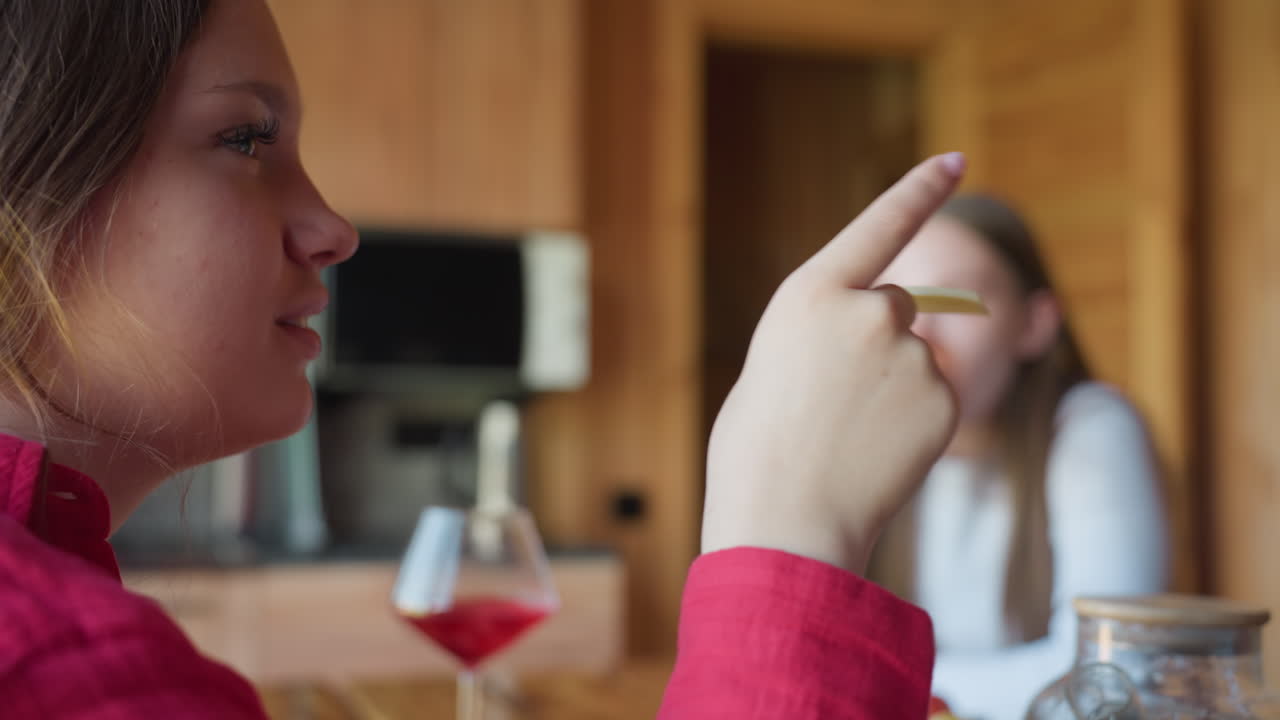 Two young women share quiet lunch moment inside warm wooden interior, one sipping red drink from glass cup while the other eats, sunlight highlights cozy mood