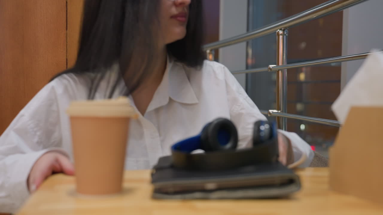 Close up of woman placing coffee cup, headphones, and tablet on wooden table before sitting down in cozy modern cafe with soft lighting and city view reflecting through large glass window