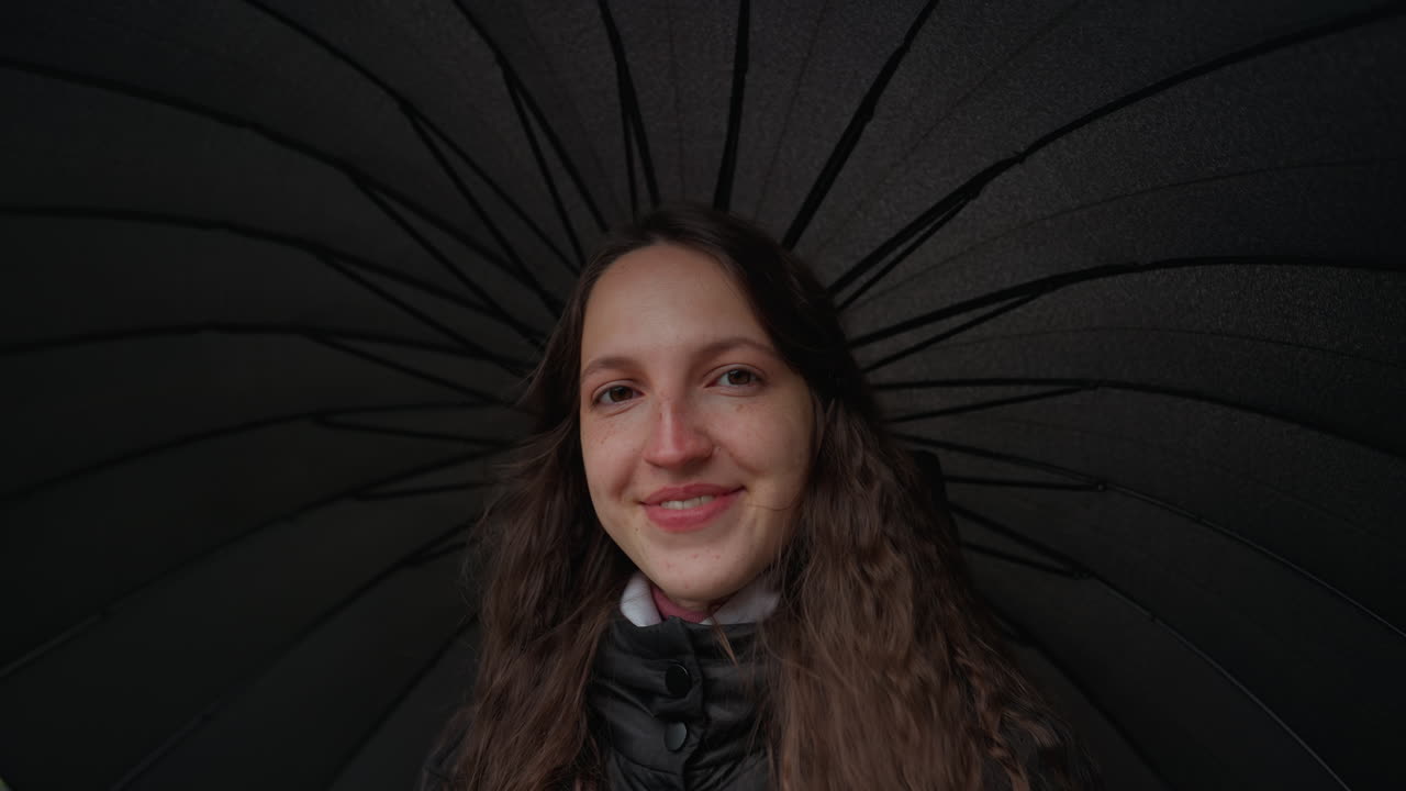 Kvinde smiler under sort paraply, nærbillede, skiftende udtryk fra neutral til grin, dråber på stoffet, hyggeligt tørklæde, blødt naturligt lys, ægte glad stemning, øjenkontakt