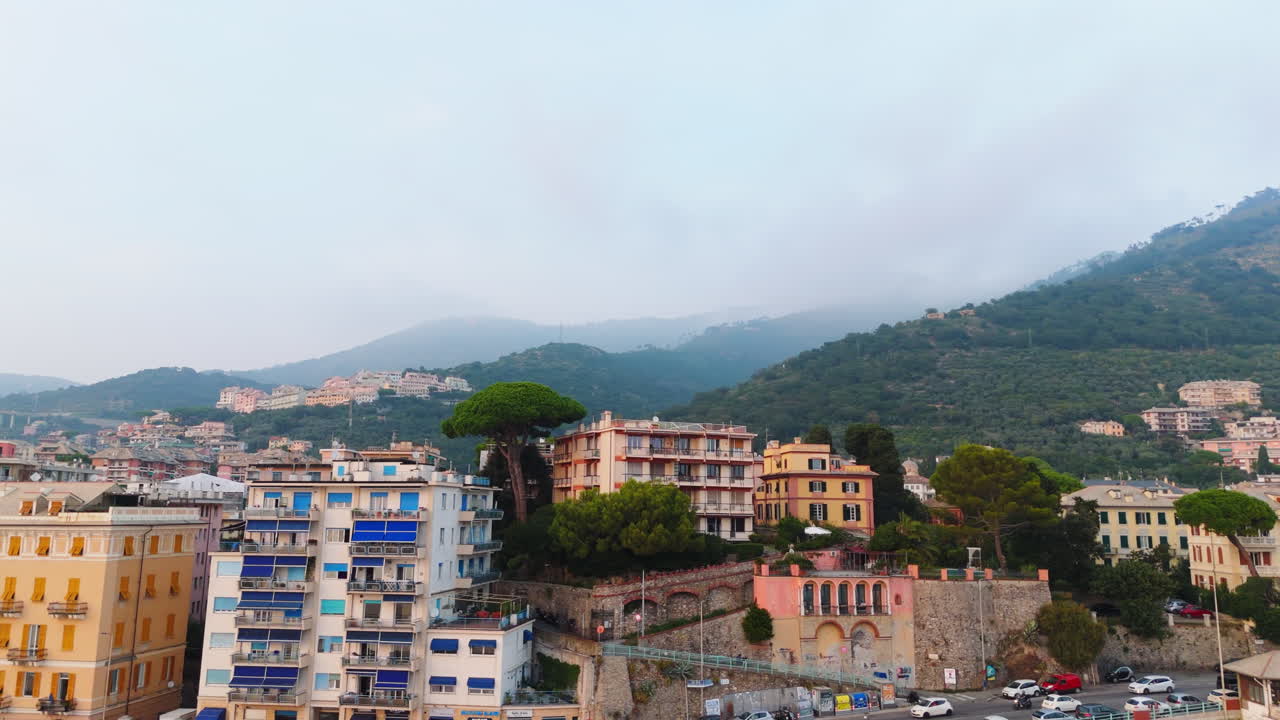 The drone performs a panning and slightly orbiting motion above colorful rooftops of a Ligurian coastal town, with green mountains in the background