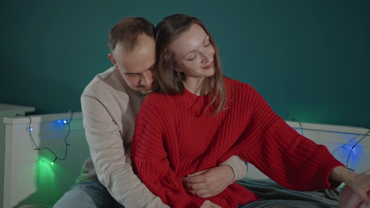 Un uomo siede dietro una donna sul divano, avvolgendole le braccia intorno alla vita mentre lei si appoggia all'indietro sorridendo e si allunga verso una coperta rosa, creando un momento domestico giocoso e accogliente in una luce calda e soffusa.