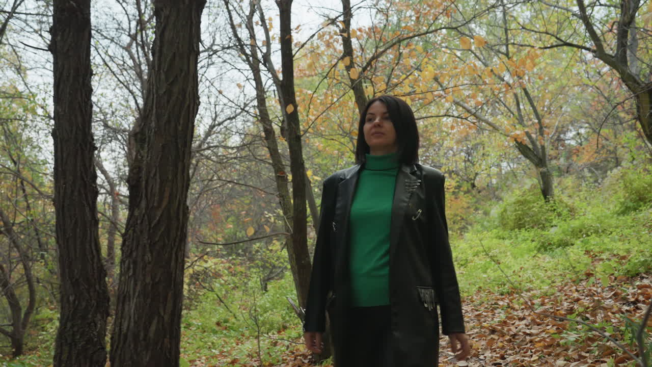 Donna con giacca di pelle, bosco autunnale. Dolcevita verde in piedi tra gli alberi con foglie cadute, cielo nuvoloso che crea un'atmosfera cinematografica suggestiva, espressione contemplativa sicura, musicista indie