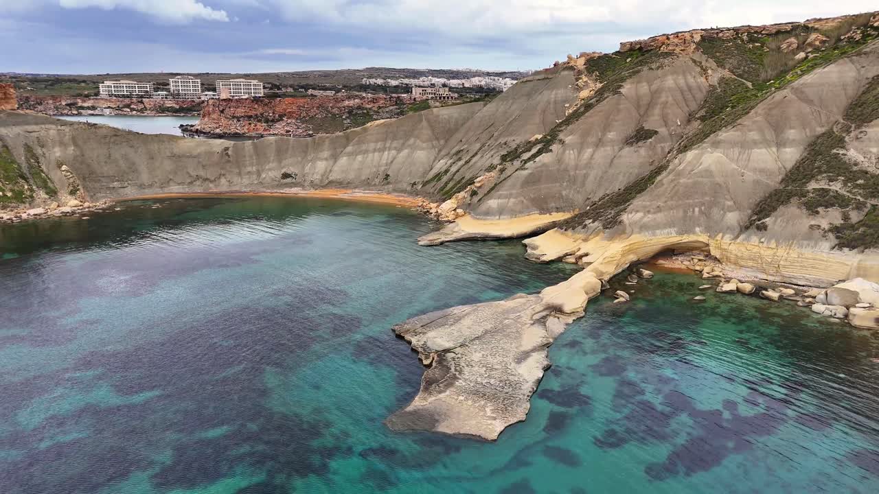 Veduta aerea con drone di ripide scogliere calcaree che si incurvano attorno all'acqua turchese nella Baia di Qarraba, Malta, con una costa rocciosa frastagliata e mare calmo alla luce del giorno.