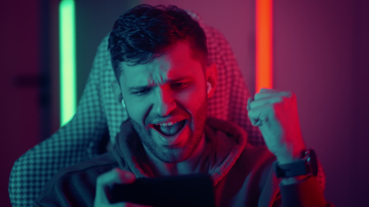A young man with glasses a professional gamer on a mobile phone plays games and emotionally rejoices in the victory. Emotional young man playing mobile games in a neon light. High quality 4k footage