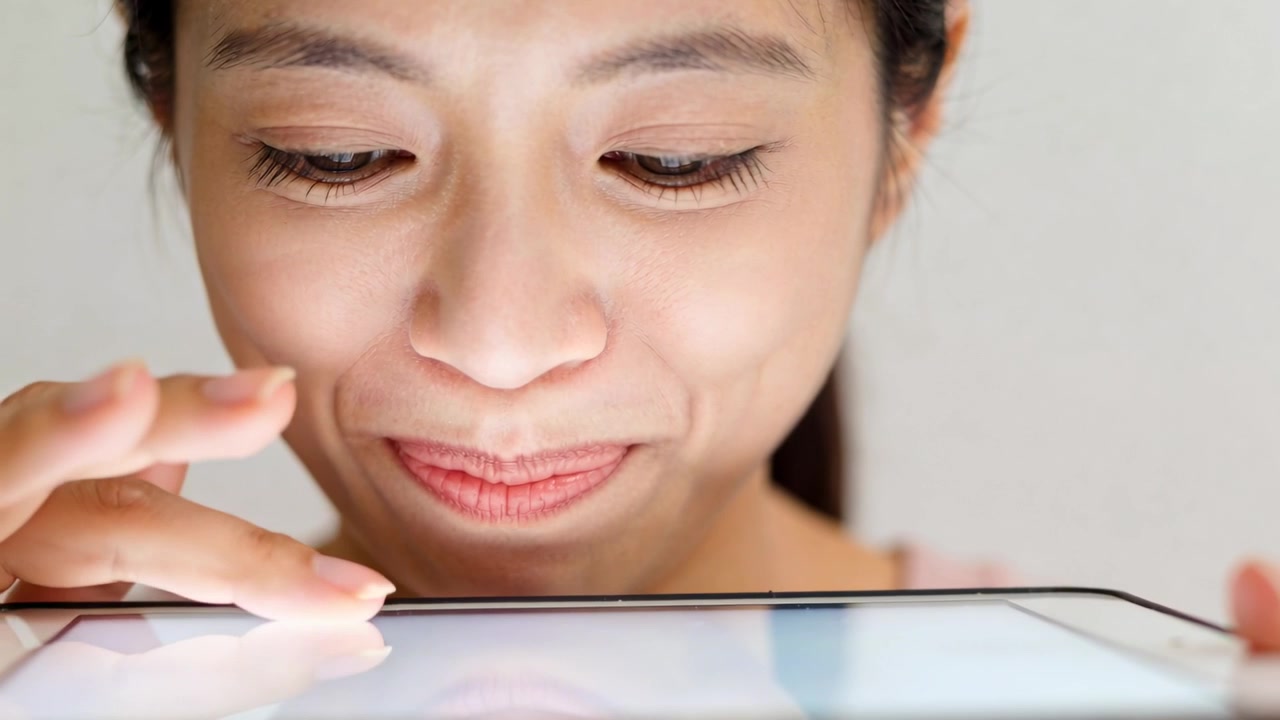 ピンクのトップスを着た女性が自宅で寄りかかりながら、顔の近くでタブレットをタップしたりスワイプしたりしており、アプリのアイコンが見えている。