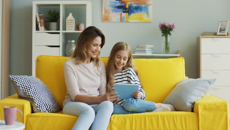 blonde mother and daughter smiling and watching something on tablet while sitting on sofa in living room