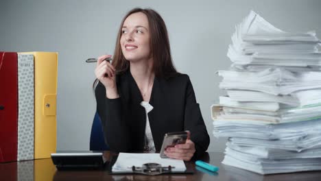 pensive businesswoman in black office uniform sitting among piles of paperwork, difficult financial calculations and overtime working day. smiling manager using smartphone and filling application blanks. data analyzing and deadline concept