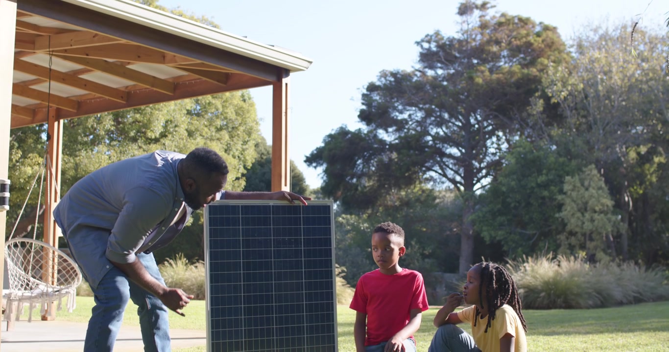 Afrikansk mand placerer solpanel på græsset ved verandaen, vinkler det mod sollys og underviser to børn.