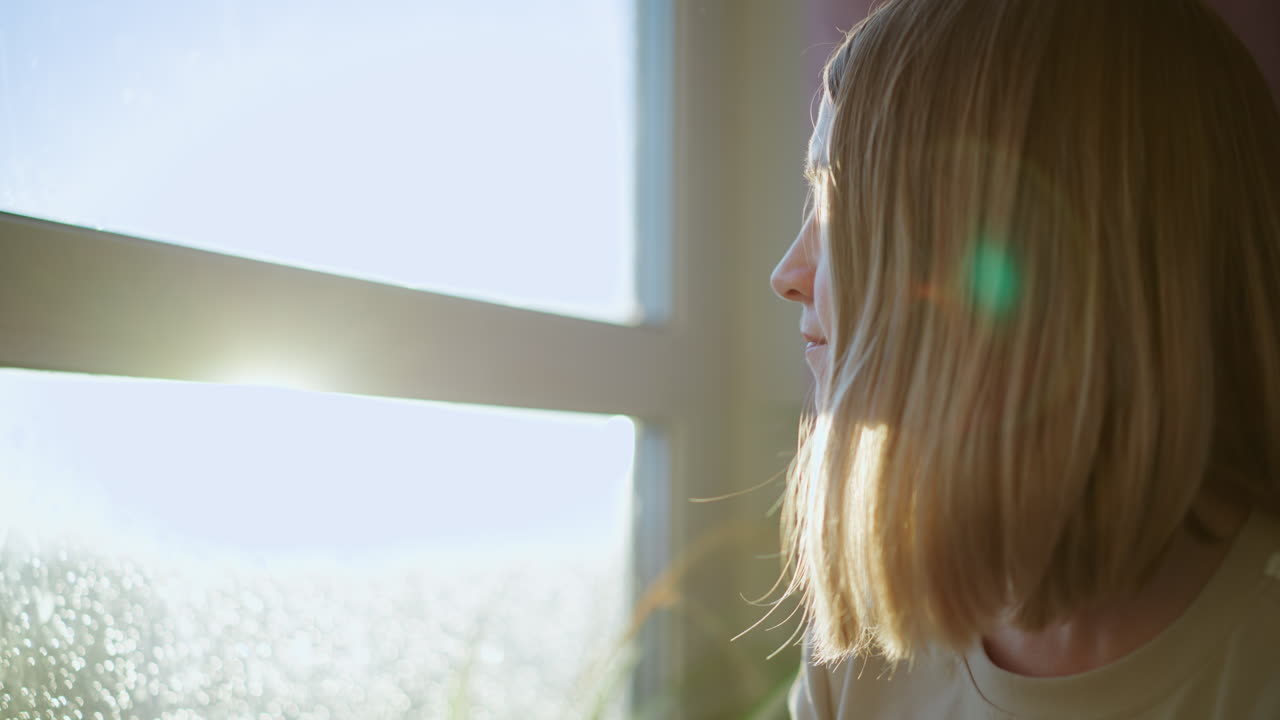 Young blonde woman sitting near window in soft sunlight, side profile view with natural glow and lens flare, peaceful moment of relaxation and contemplation indoors, serene lifestyle atmosphere