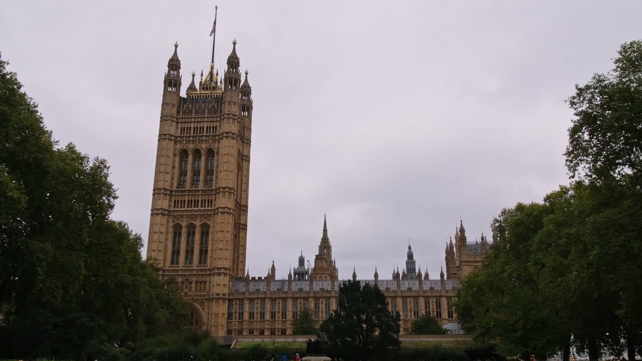 People walk past the Victoria Tower Palace Of Westminster in the Tower Gardens South Park In London, United Kingdom. Low wide shot.