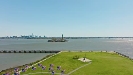 Aerial-approaching-shot-of-Ellis-Island-with-waving-american-flags,Statue-of-Liberty-and-Skyline-of-New-York-city-in-Background